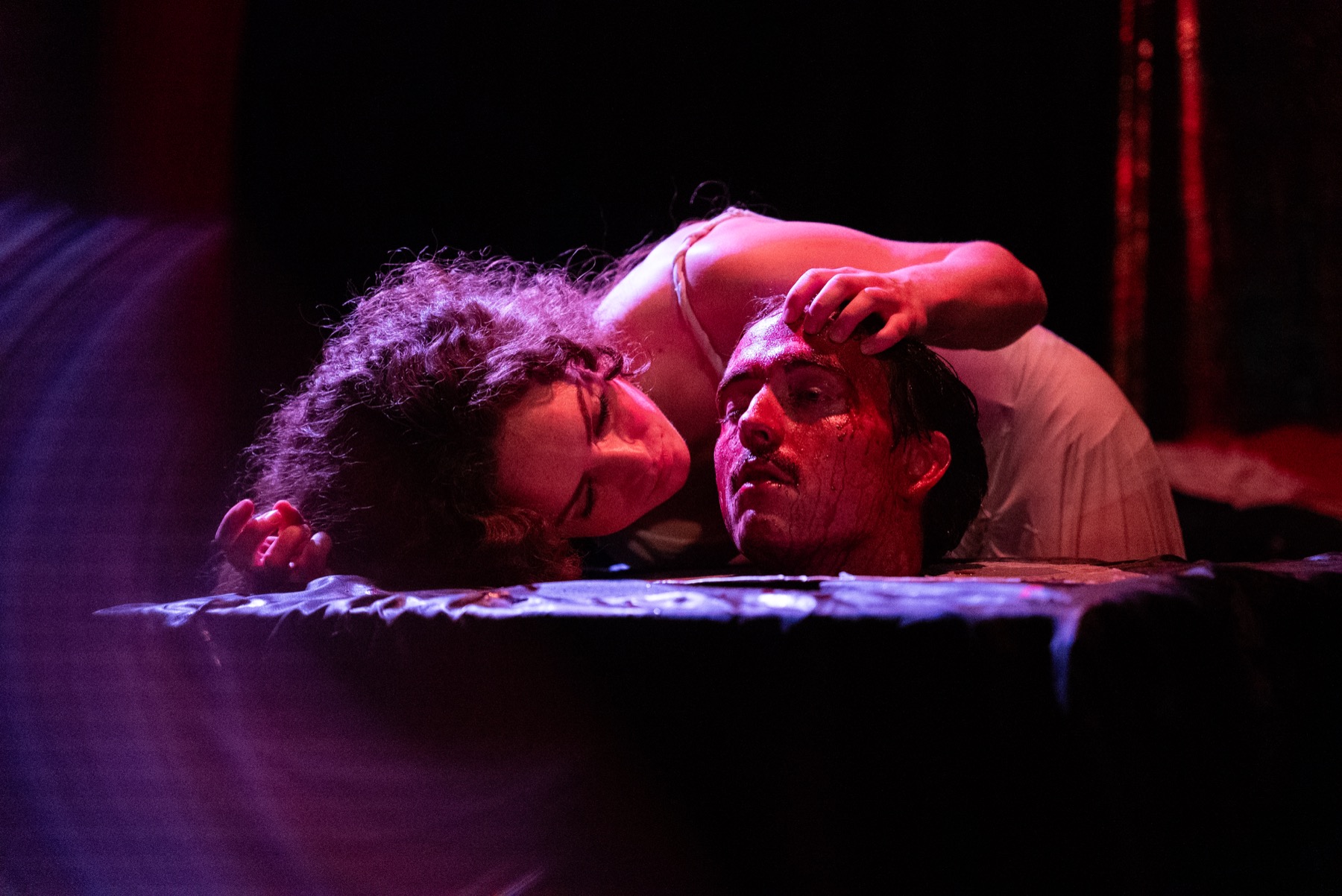 Salome leans over the head of Yokanaan under red light in DECAPITATRIX