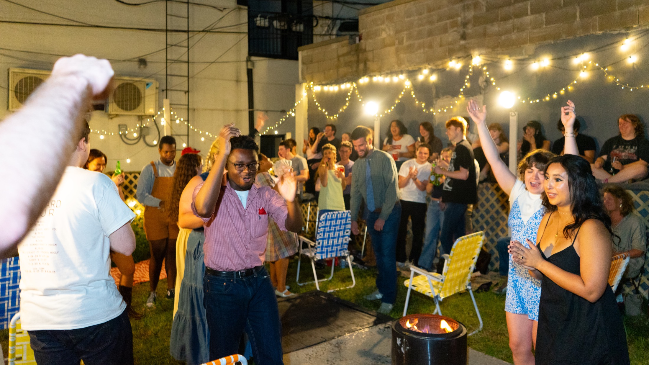 Audience and performers gathered around a backyard fire pit under string lights