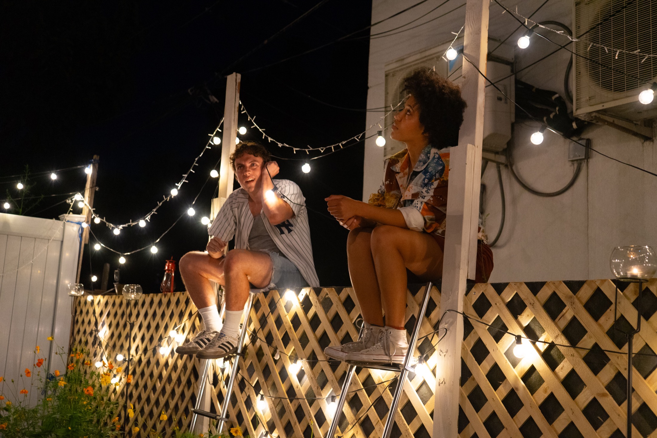 Two performers sit elevated among string lights during Our Town