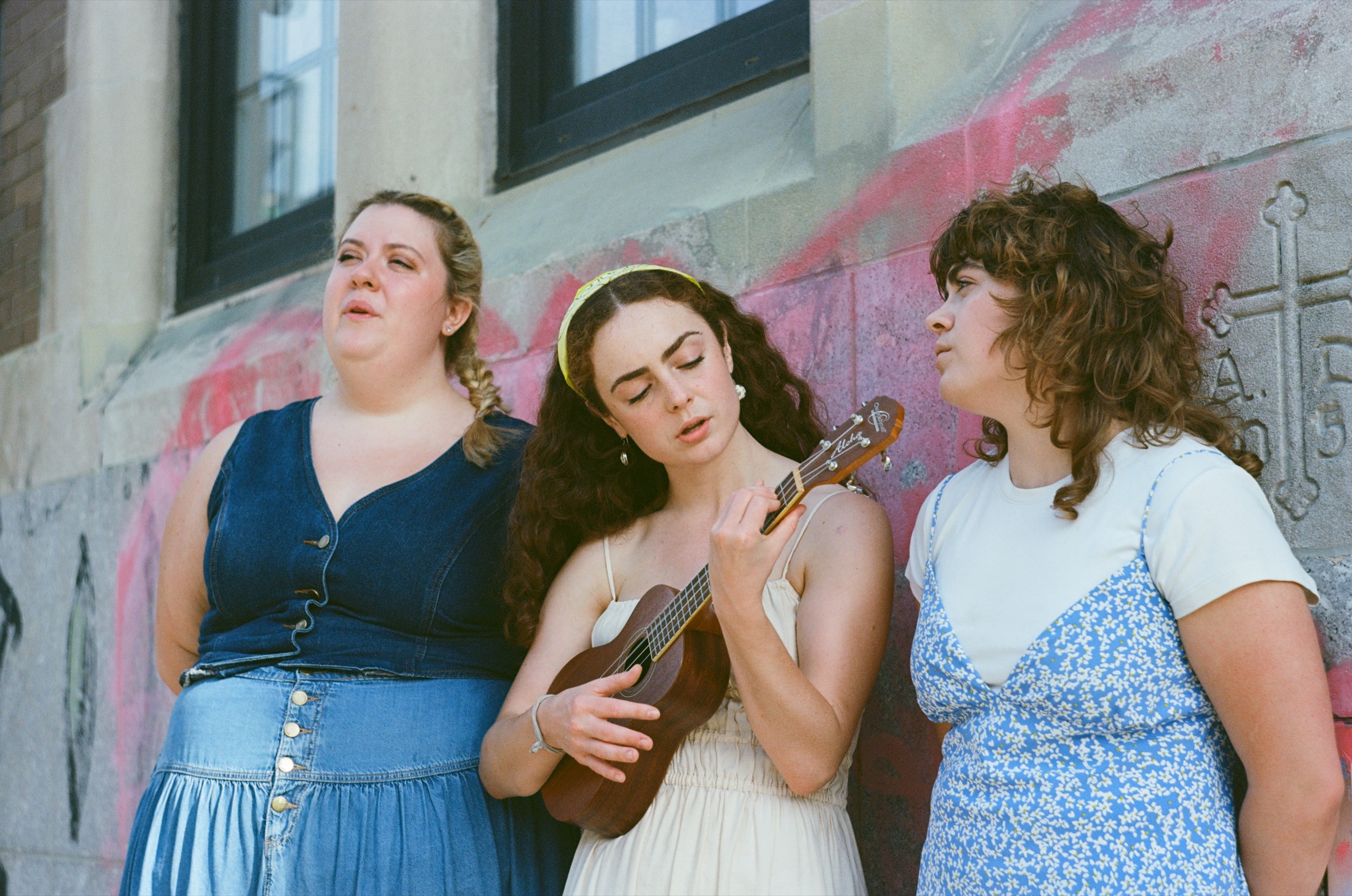 Three performers sit with a ukulele against a painted sidewalk wall
