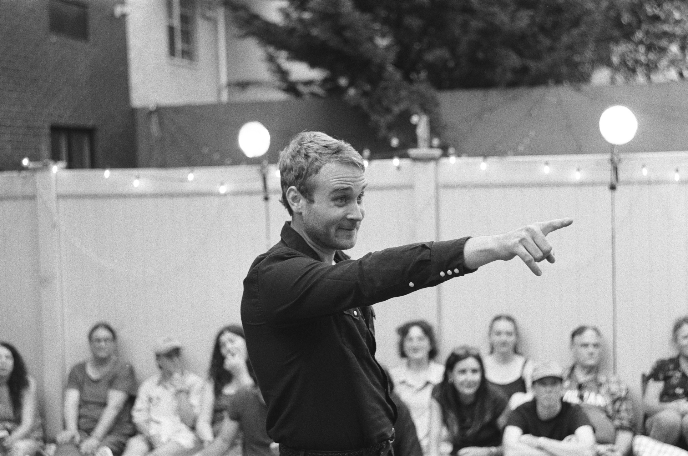 The Stage Manager speaks under glowing string lights in black and white