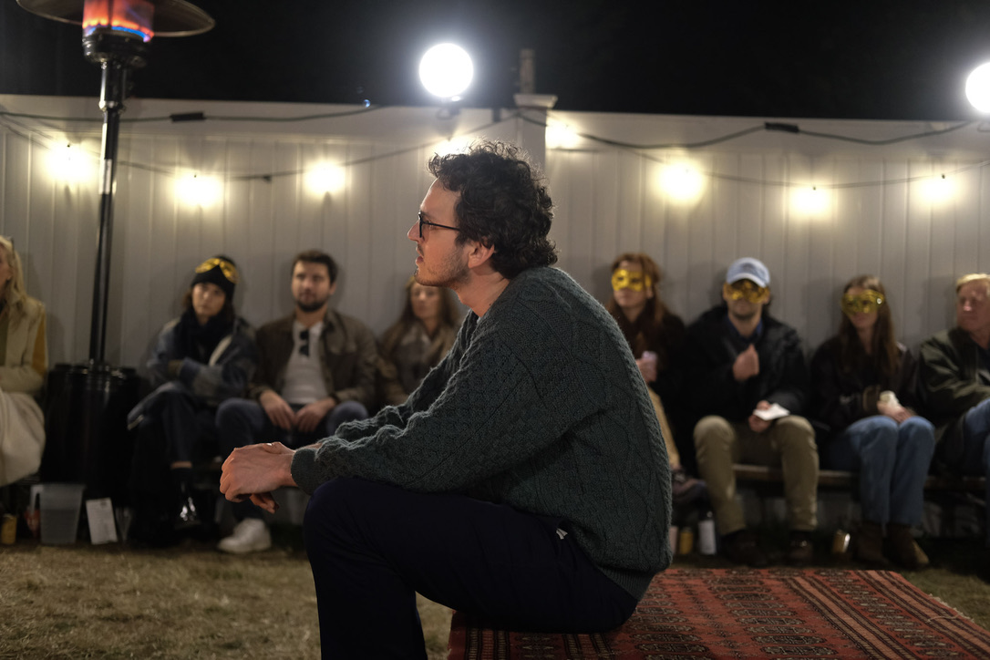 A performer sits on a rug before masked audience members