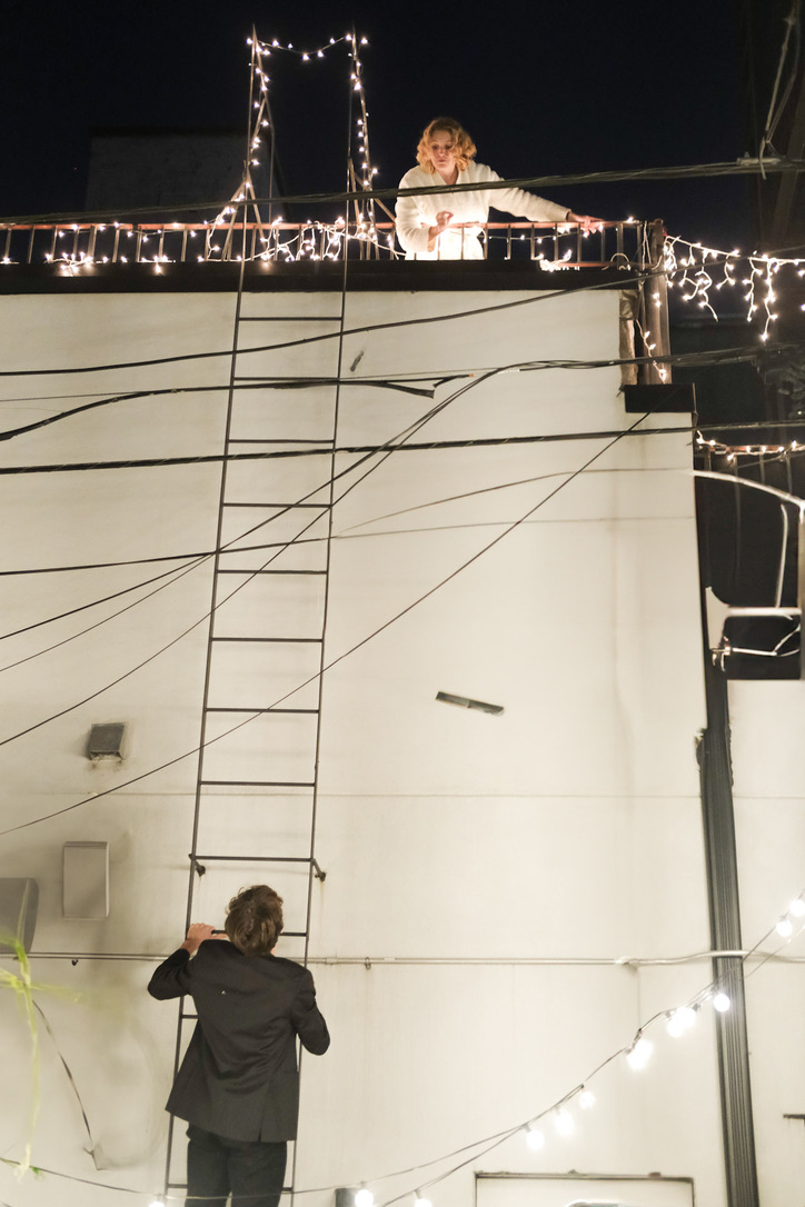 Juliet on a rooftop balcony above Romeo with string lights and a ladder