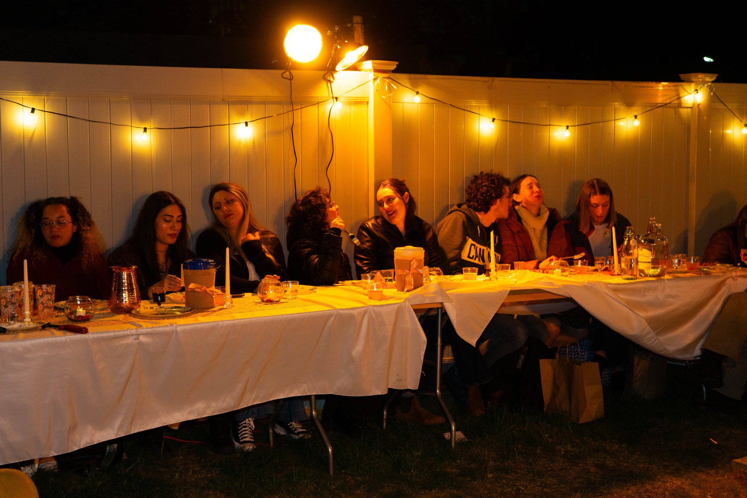 Audience members seated at a long backyard dinner table under warm string lights