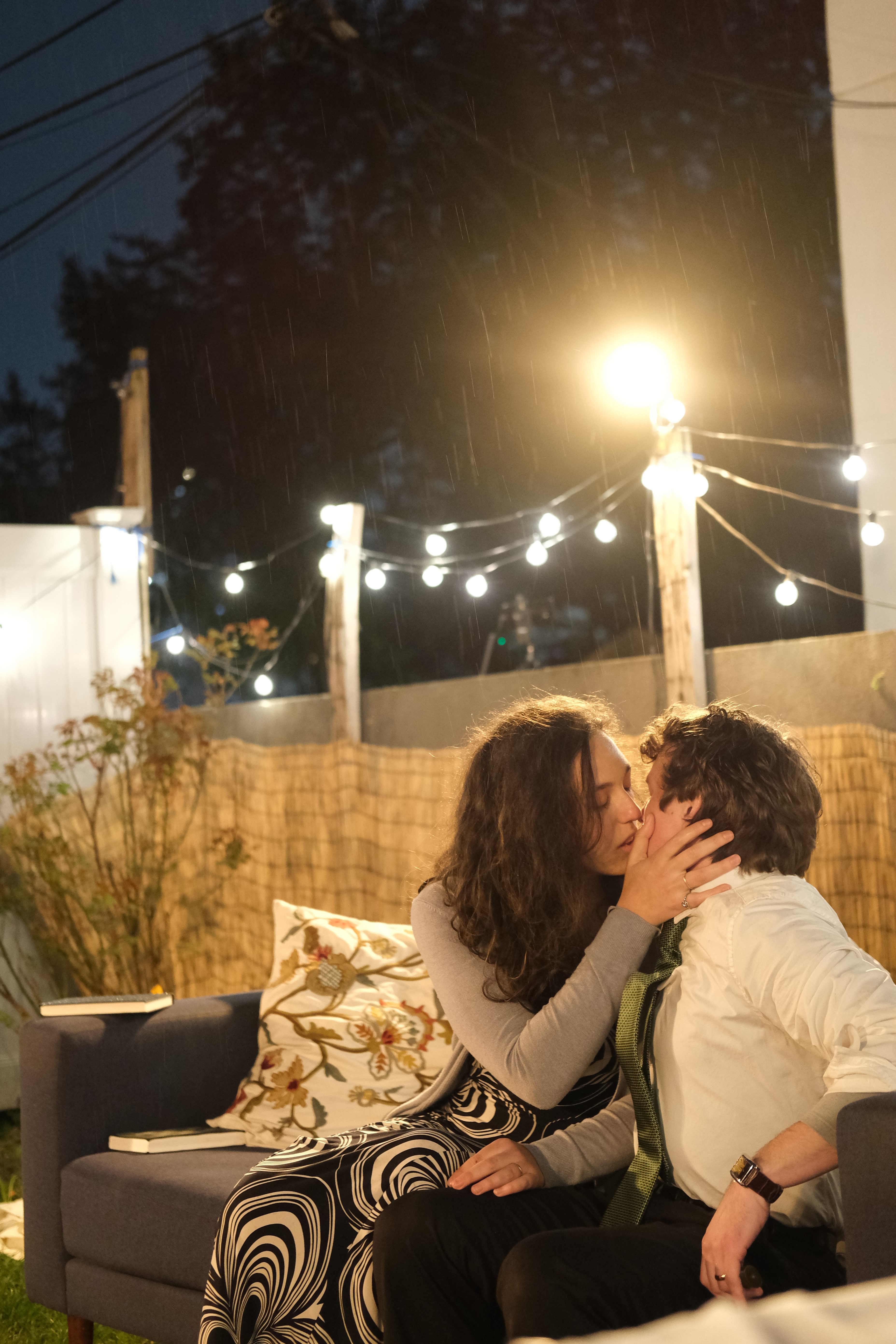 Nick and Honey kiss on an outdoor couch under string lights and rain