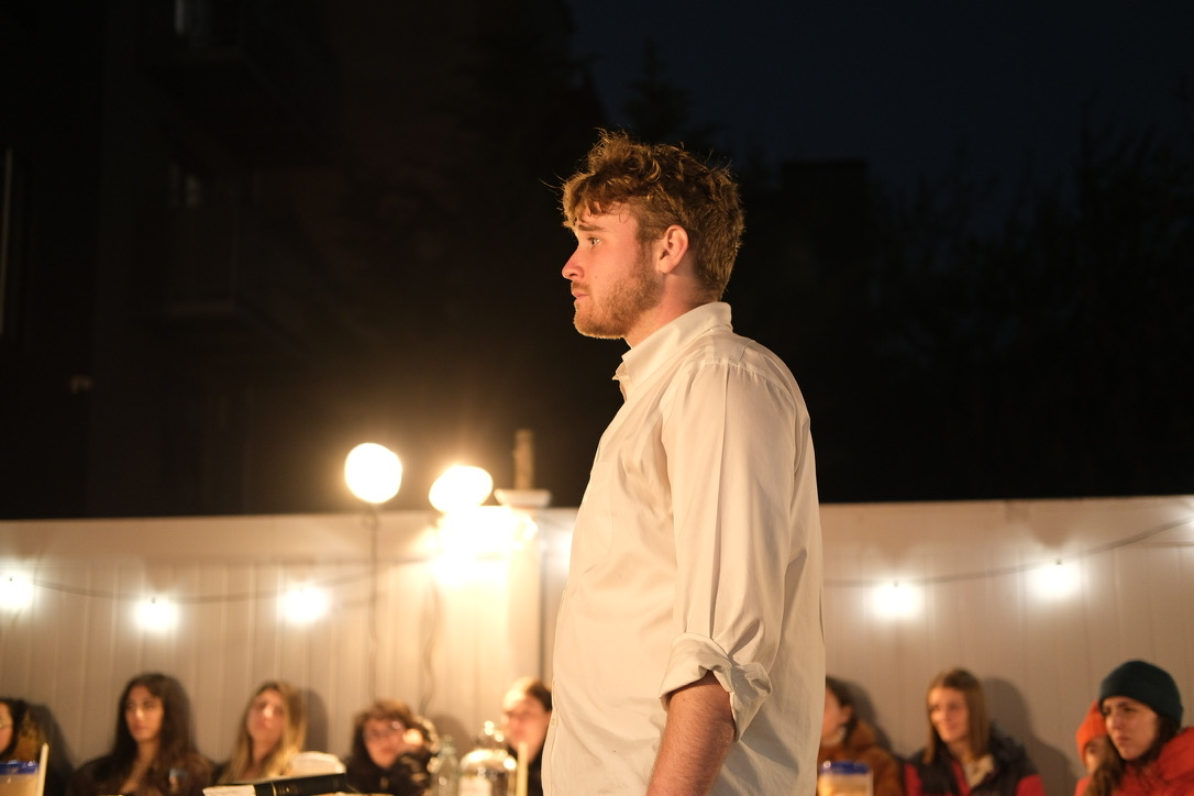 Nick stands under backyard lights while audience members look on